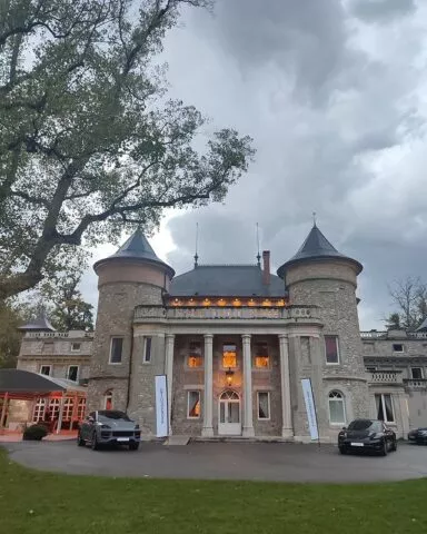 Un grand manoir en pierre avec deux tours cylindriques, chacune surmontée d'un toit conique, se dresse sous un ciel nuageux. Les fenêtres éclairées suggèrent le soir. Deux voitures de luxe sont garées devant. Un grand arbre encadre le côté gauche et des bannières flanquent l'entrée principale. L'herbe entoure la zone.